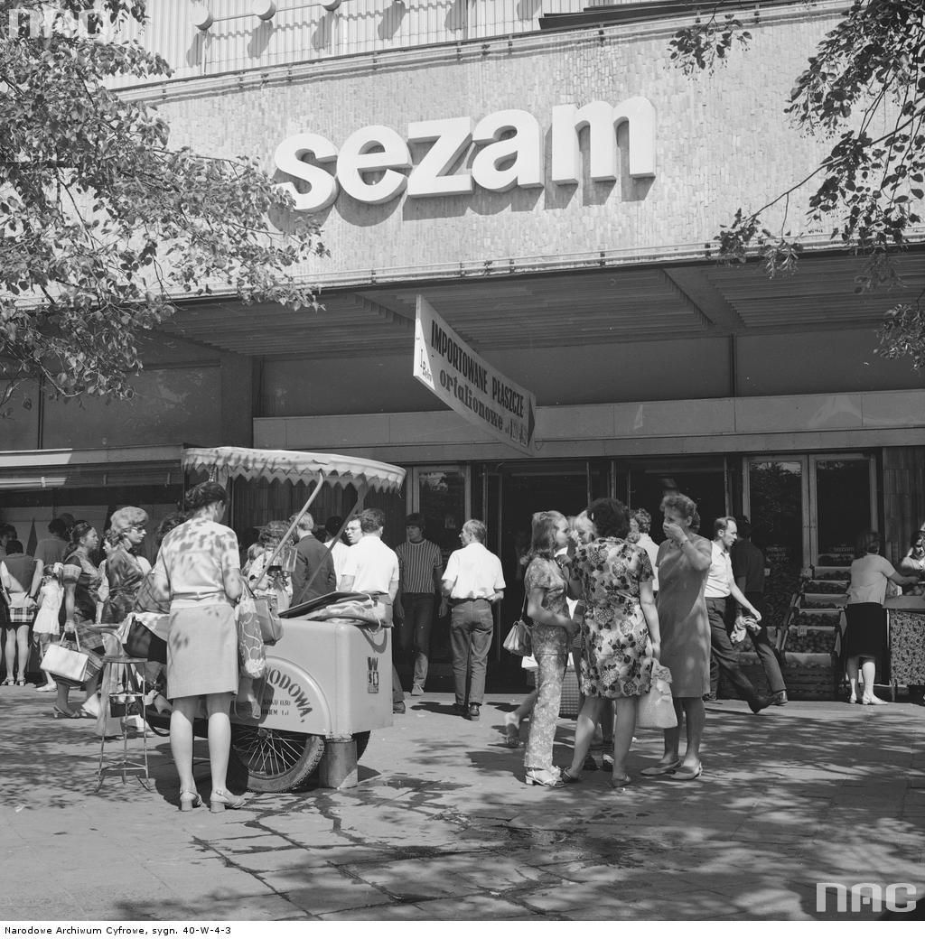 Przechodnie przed Domem Towarowym "Sezam". Na pierwszym planie saturator z wodą sodową. Widoczny transparent reklamowy "Importowane płaszcze ortalionowe". Fot. Narodowe Archiwum Cyfrowe