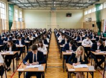 Maturzyści ubrani na galowo piszący egzamin dojrzałości na dużej sali