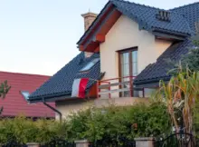Nowoczesny prywatny dom z ciemnym dachówką, balkon i flagą Polski, kamiennym ogrodzeniem i zielonymi drzewami w dzielnicy mieszkalnej.