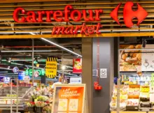 Podświetlany znak Carrefour zdobi nowoczesne wejście do sklepu. Wewnątrz widać świeże jedzenie, owoce i kwiaty.